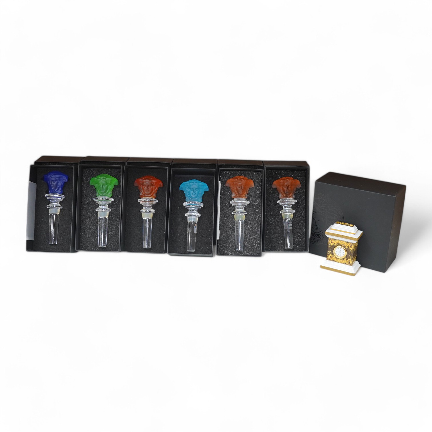 Six boxed Rosenthal for Versace glass Medusa head wine bottle stoppers in various colours and a boxed miniature timepiece, 8.5cm high (7). Condition - good.